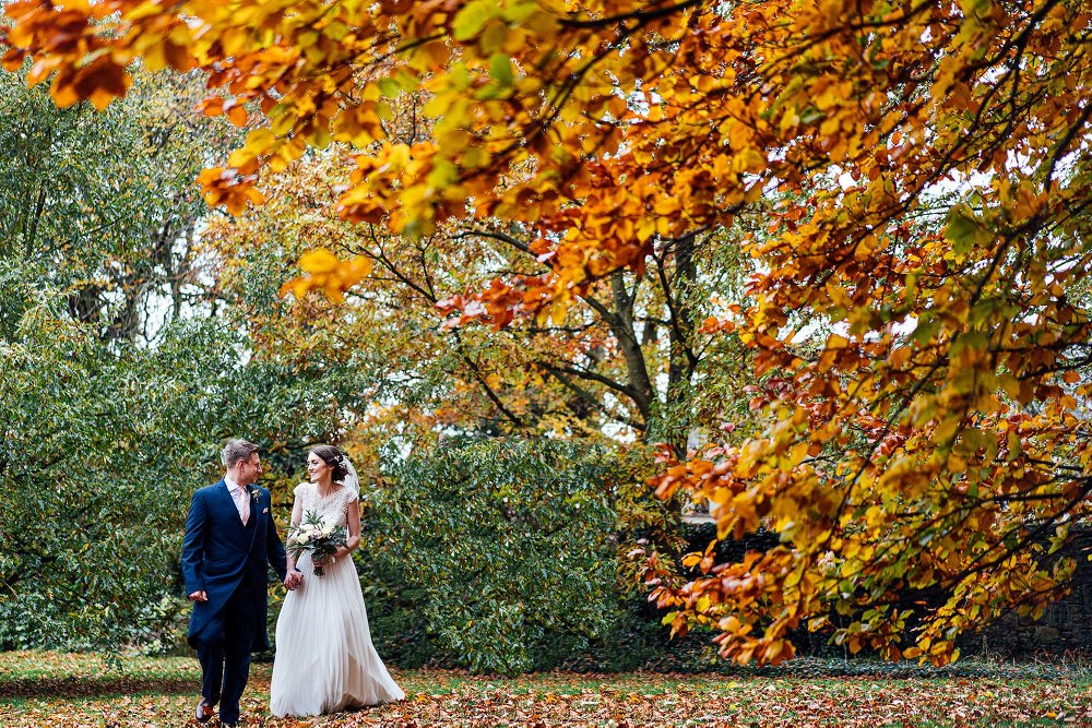 Bride & Groom walking in grounds at West Tower with orange leaves on the trees at their autumn wedding