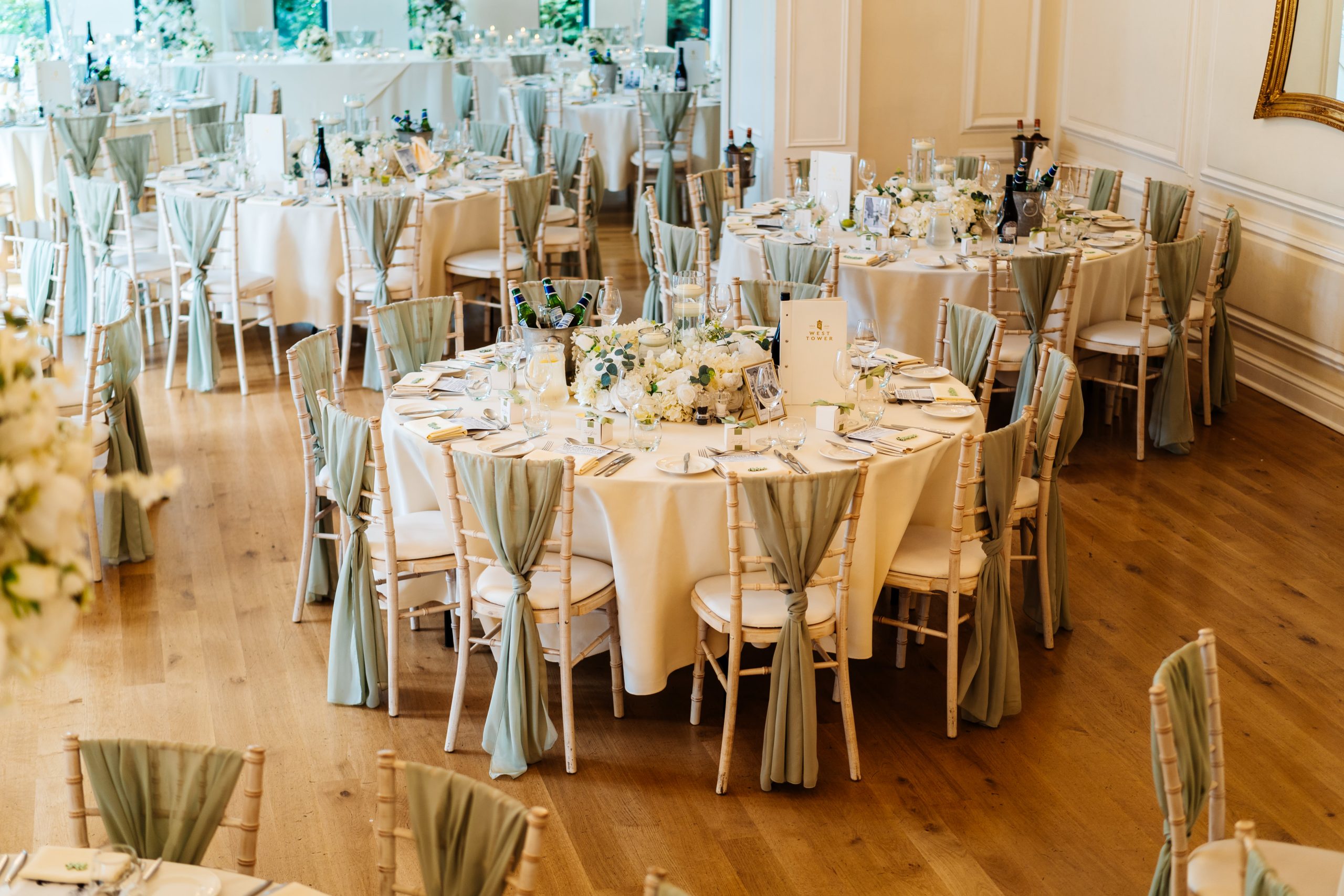 A wedding breakfast set up with round tables surrounded by sage green colour sashes on Chiavari chairs. Each setting is laid out with a napkin, side plate, wine glass, water glass and cutlery. This is set up in the west tower ballroom who are attending the wedding show