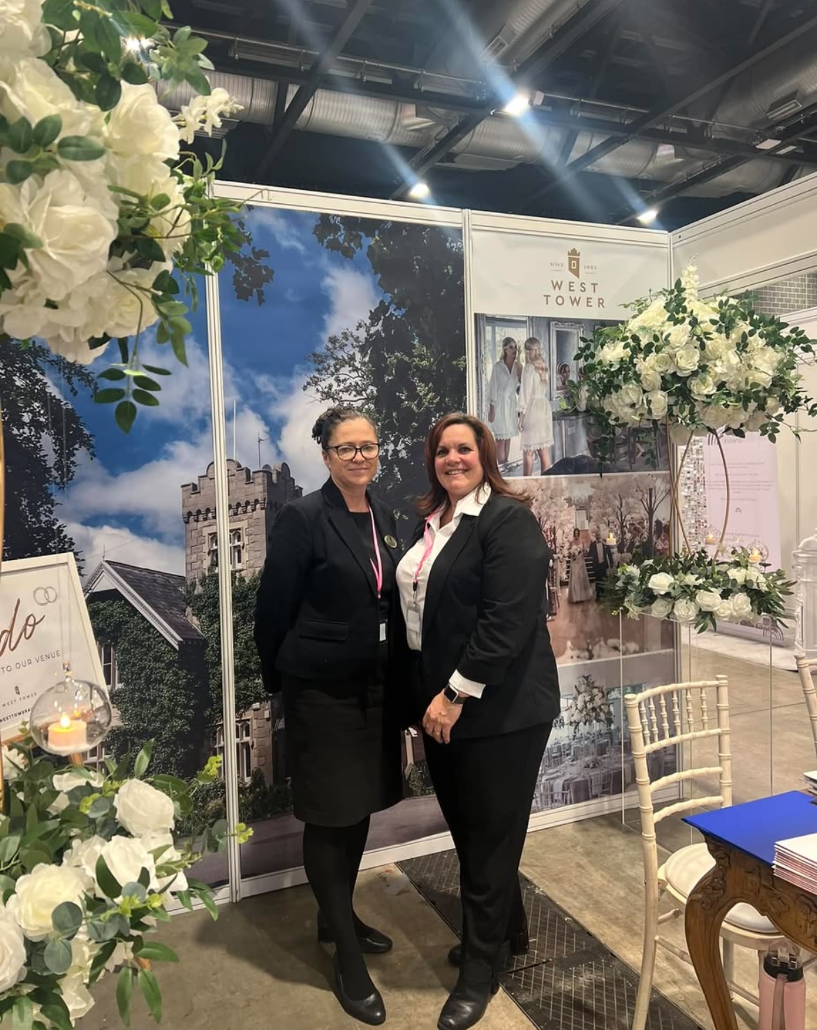 Two west tower staff standing facing the camera smiling at an exhibition stand for west tower in front of a picture of west tower main house with flower arrangements hung up next to them