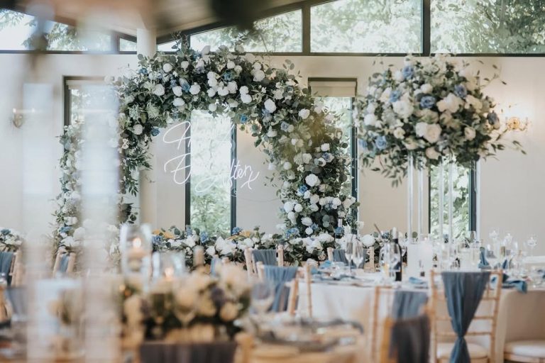 Ballroom at West Tower styled with a large white and blue floral arch, tall flower centrepieces and chairs dressed with blue sashes for a wedding breakfast.