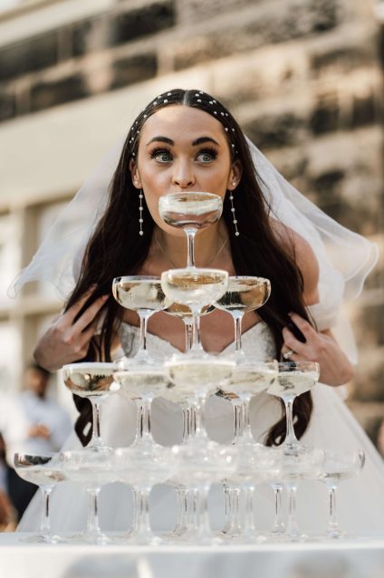 Smiling bride leaning in to sip from the top glass of a champagne tower during her wedding reception at West Tower.