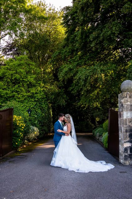 Bride and groom standing together in the driveway between the stone entrance gates with West Tower signs and trees behind them.