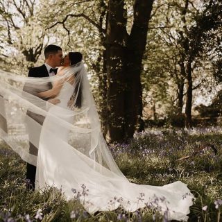 Bride and groom kissing in a woodland clearing at West Tower, surrounded by bluebells with the bride’s long veil flowing around them.