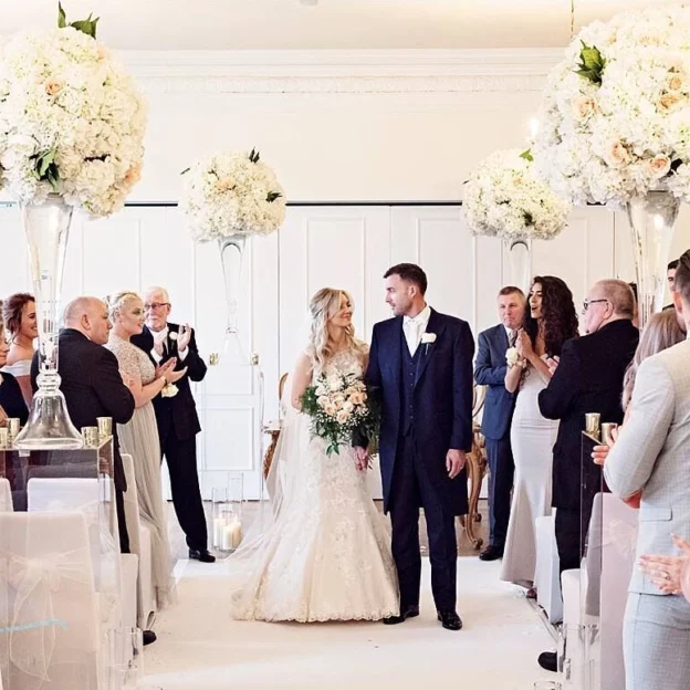 Bride and groom walking back down the aisle in West Tower’s ceremony room, surrounded by guests clapping beneath tall white flower arrangements.