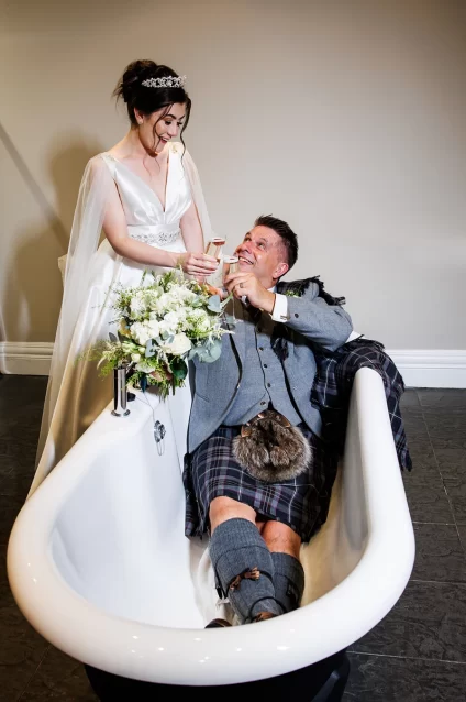 Bride standing beside a freestanding bathtub while the groom sits in the tub, both laughing and toasting with champagne at West Tower.