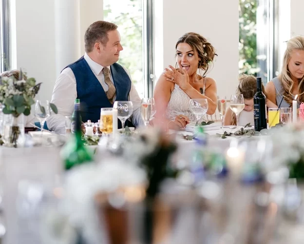 Bride smiling during the wedding speeches at West Tower, sitting at the top table with guests and glasses raised around her.