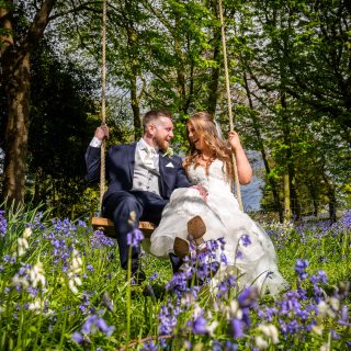 A bride and groom sat facing each other smiling on the swing in the west tower woodland walk surrounded by blue and white flowers in the grass